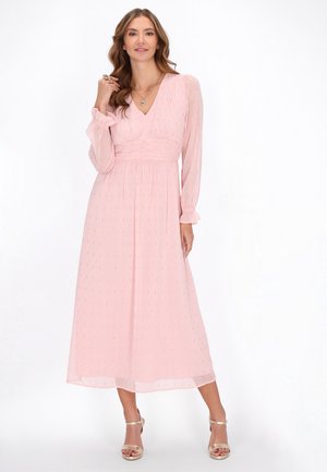 Maxikleid aus hellrosa Chiffon mit V-Ausschnitt, langen Ärmeln, gerafftem Bund und subtilen goldenen Punkten. Kombiniert mit metallischen Sandalen.