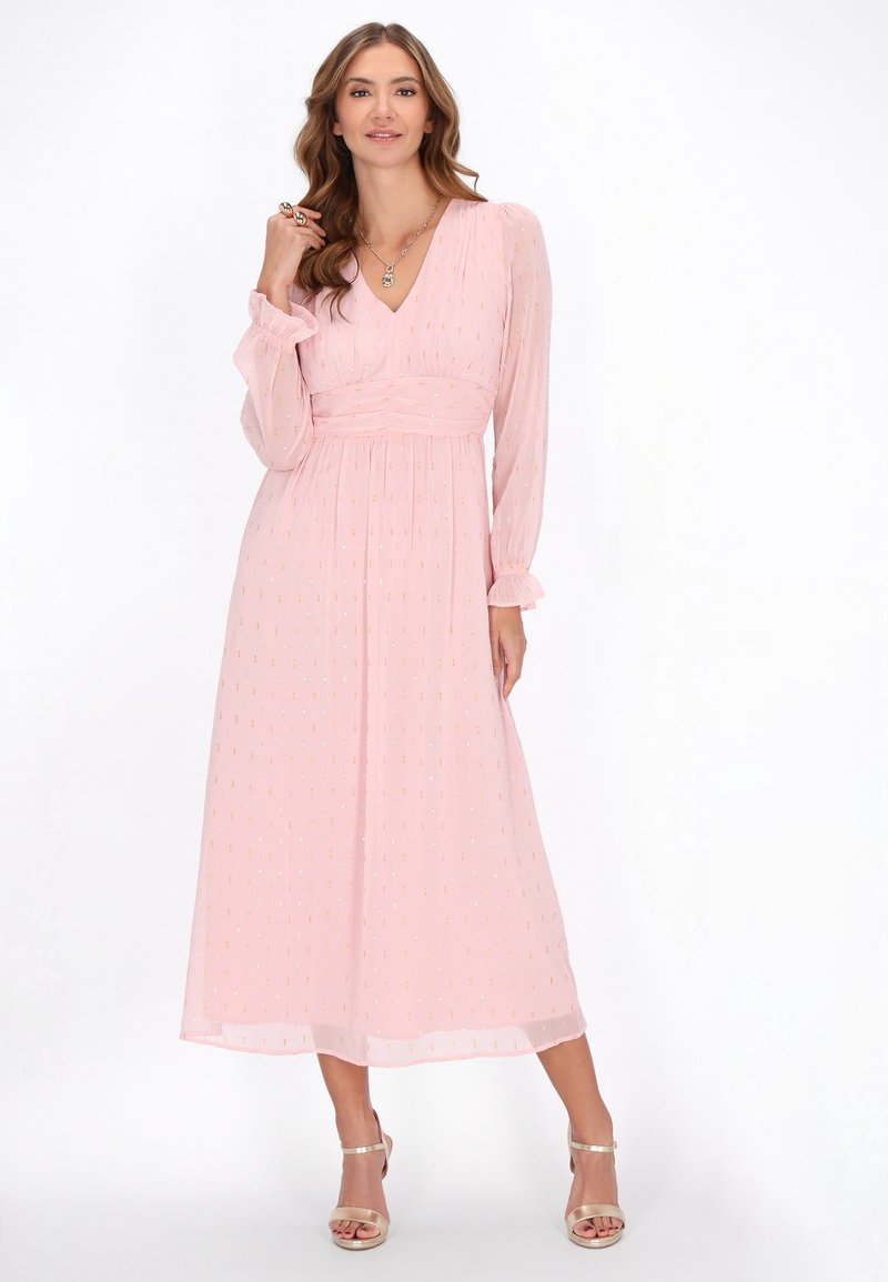 Rochie maxi din chiffon roz deschis cu decolteu în V, mâneci lungi, talie strânsă și un model subtil cu puncte aurii. Asortată cu sandale metalice.