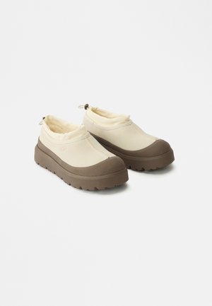 Μπεζ καμβάς παπούτσια slip-on με καφέ καουτσούκ σόλες, διαθέτοντας υφασμάτινη μύτη και ελαστική περιφέρεια για εύκολη εφαρμογή.