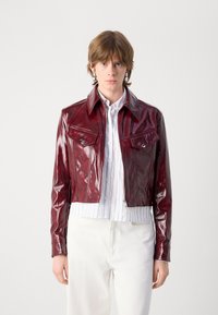 Burgundy leather cropped jacket with a glossy finish, featuring chest pockets, a pointed collar, and silver button accents. Worn over a striped shirt.