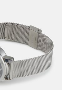 Correa de reloj de malla metálica en plata, con una textura suave y un cierre ajustable. Grabada con "acero inoxidable" en el hardware.