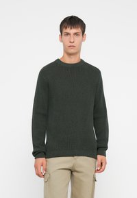 Junger Mann, der nach vorne schaut und einen dunkelgrünen gerippten Strickpullover sowie beigefarbene Cargo-Hosen trägt, vor einem schlichten weißen Hintergrund.