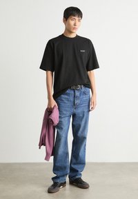 T-shirt negru din bumbac cu un logo pe față, asociat cu blugi din denim albastru. O pulover violet este ținut într-o mână, stilizat cu pantofi negri.