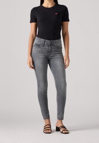 Camiseta negra ajustada con un pequeño logo rojo, combinada con unos pantalones ajustados grises de cintura alta. Modelo con sandalias de tacón bloque negras.