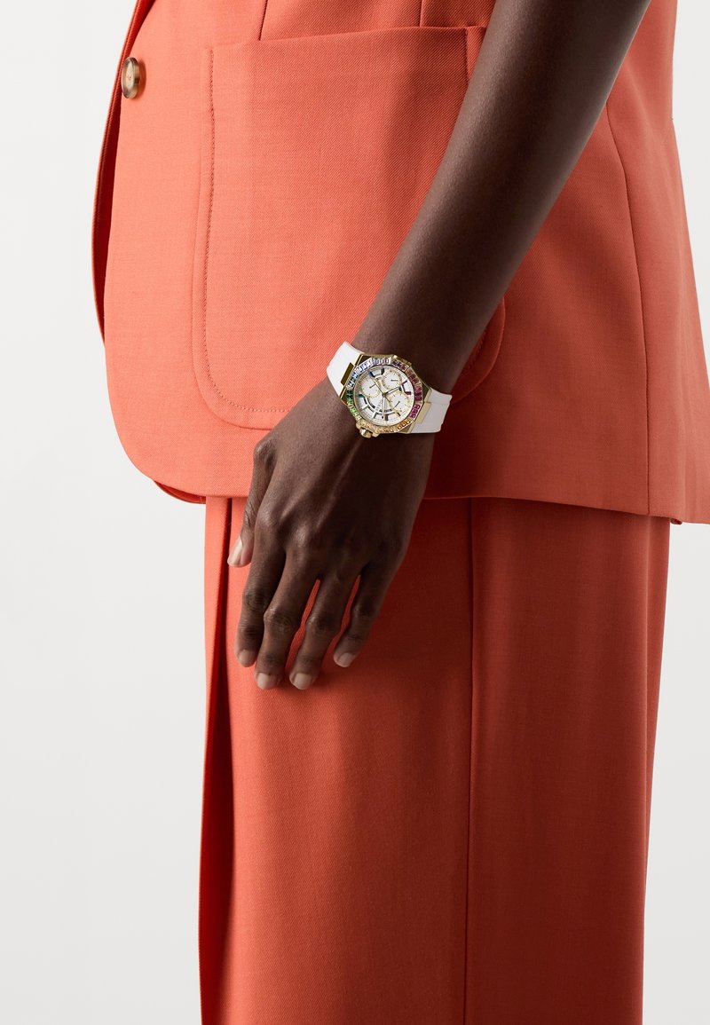 Personne à la peau foncée portant un costume orange avec une montre-bracelet blanche dotée d'une lunette colorée et de détails dorés à son poignet gauche.