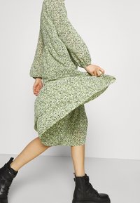 Vihreä kukkamekko pitkillä hihoilla, kirjattu vyötärö ja virtaava helma, yhdistettynä mustiin nilkkureihin kontrastiksi.