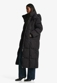 Geacă lungă neagră de tip puf, cu glugă, având un design matlasat, închidere cu fermoar și capse. Poartă peste blugi evazați închiși la culoare și pantofi negri cu toc.