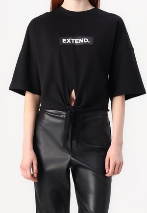 Czarny oversize'owy T-shirt z napisem "EXTEND.", związany na supeł z przodu, noszony z wysokim stanem czarnych skórzanych spodni na stojącej osobie.