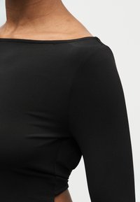 Κοντινό πλάνο ενός ατόμου που φοράει εφαρμοστό μαύρο μακρυμάνικο τοπ με φαρδιά λαιμόκοψη που αφήνει ακάλυπτο τον ώμο και την περιοχή πάνω από το στήθος.