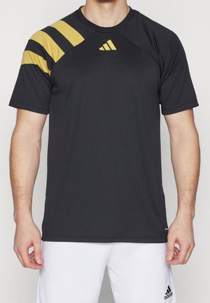 Homme portant un maillot de sport Adidas noir avec trois bandes diagonales jaunes sur l'épaule gauche et un short blanc Adidas.