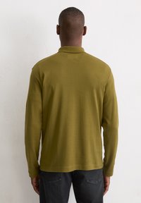 Langärmliges olivgrünes Polohemd aus weichem Stoff, mit geripptem Kragen und geradem Saum, kombiniert mit dunklen Jeans.