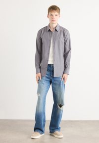 Camisa de botones gris a rayas, camiseta blanca interior, jeans claros desgastados y zapatillas beiges, de pie contra un fondo neutro.