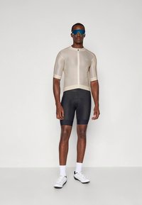 Kolesarski dres v bež barvi s kratkimi rokavi in zadrgo spredaj. Črne kratke hlače z elastičnim prileganjem. Bele superge s črnimi poudarki in modna modra sončna očala.