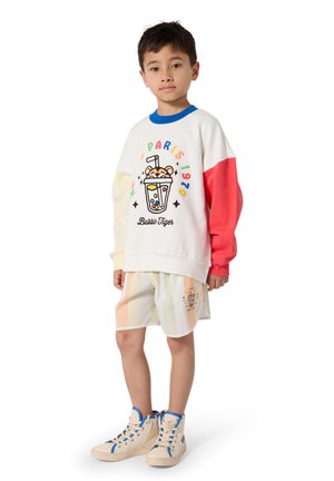 Chłopiec w kolorowej bluzie z długim rękawem z grafiką bubble tea oraz w pasiastych szortach, stojący w beżowych wysokich trampkach.