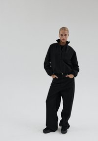 Kobieta stoi z rękami w kieszeniach, ubrana w czarną bluzę na zamek i szerokie czarne spodnie na tle jednolitego jasnego tła.