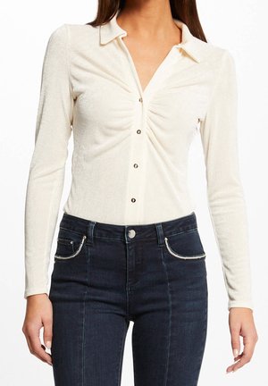 Femme portant un haut crème à manches longues boutonné avec un devant froncé et un jean sombre taille haute avec des poches bordées d'argent.