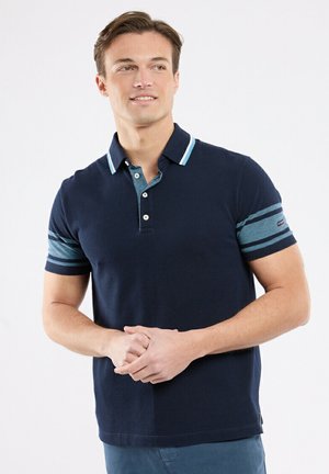 Homme portant un polo bleu marine avec des manches rayées bleu clair, debout les mains légèrement croisées, regardant sur le côté.