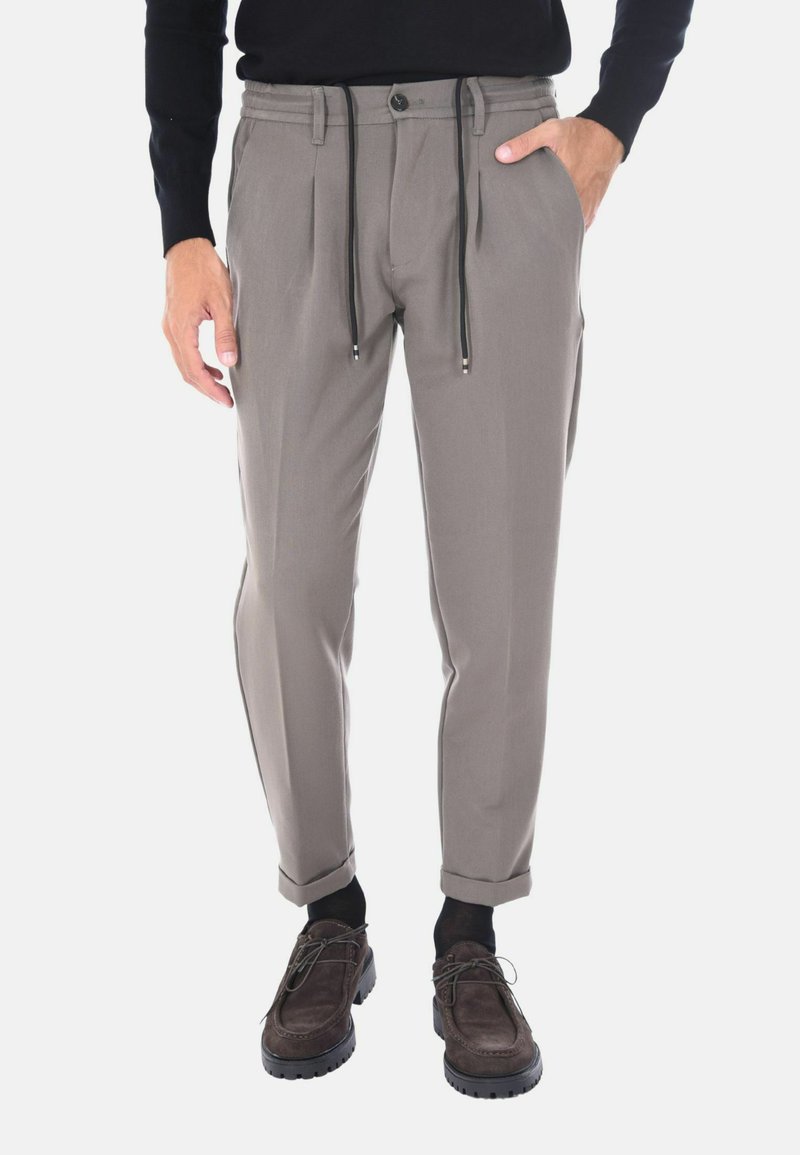 Pantaloni grigi chiari su misura con vita regolabile tramite cordoncino, dettagli con pieghe sul davanti, caviglie con risvolto e tessuto leggero; abbinati a scarpe marroni.