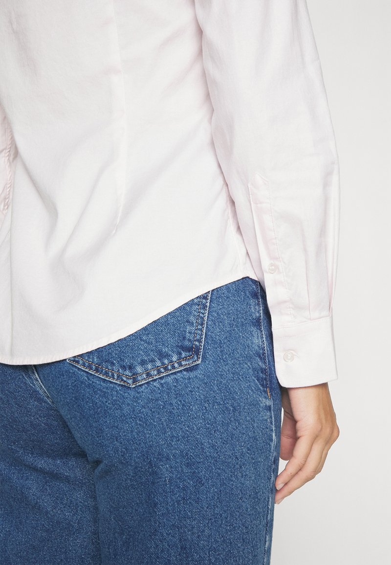 Chemise rose pâle avec un ourlet légèrement arrondi, des poignets à boutons et une texture douce, associée à un jean en denim bleu avec une poche arrière.