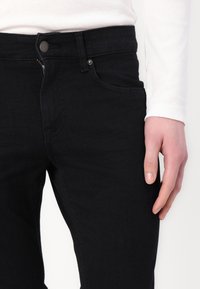 Primer plano de una persona con jeans negros y una cremallera parcialmente abierta, llevando una camisa blanca de manga larga, mostrando su mano derecha descansando a un lado.
