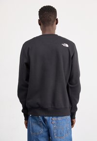 Svart hettegenser med ribbede mansjetter og hem, med en liten hvit logo på øvre rygg. Brukt sammen med blå denimjeans.