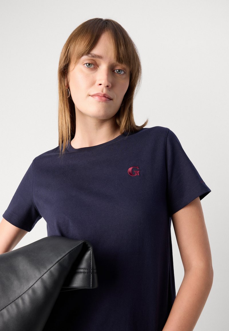 Marineblaue Baumwoll-T-Shirt mit kurzen Ärmeln, das ein kleines gesticktes rotes Logo auf der Brust trägt. Glatte Textur, minimalistisches Design.