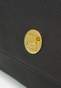 Borsa nera in pelle tessurizzata con una chiusura circolare dorata sulla quale è incisa la marca "Coccinelle", che sottolinea il lusso e la durabilità.