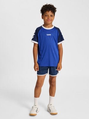 Jongen staat en lacht, draagt blauwe en marineblauwe sportkleding met witte sokken en witte sneakers, effen lichte achtergrond.