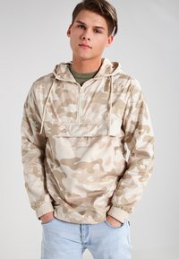 Chaqueta de media cremallera de camuflaje beige con capucha, puños elásticos y un bolsillo frontal; textura suave y material ligero.