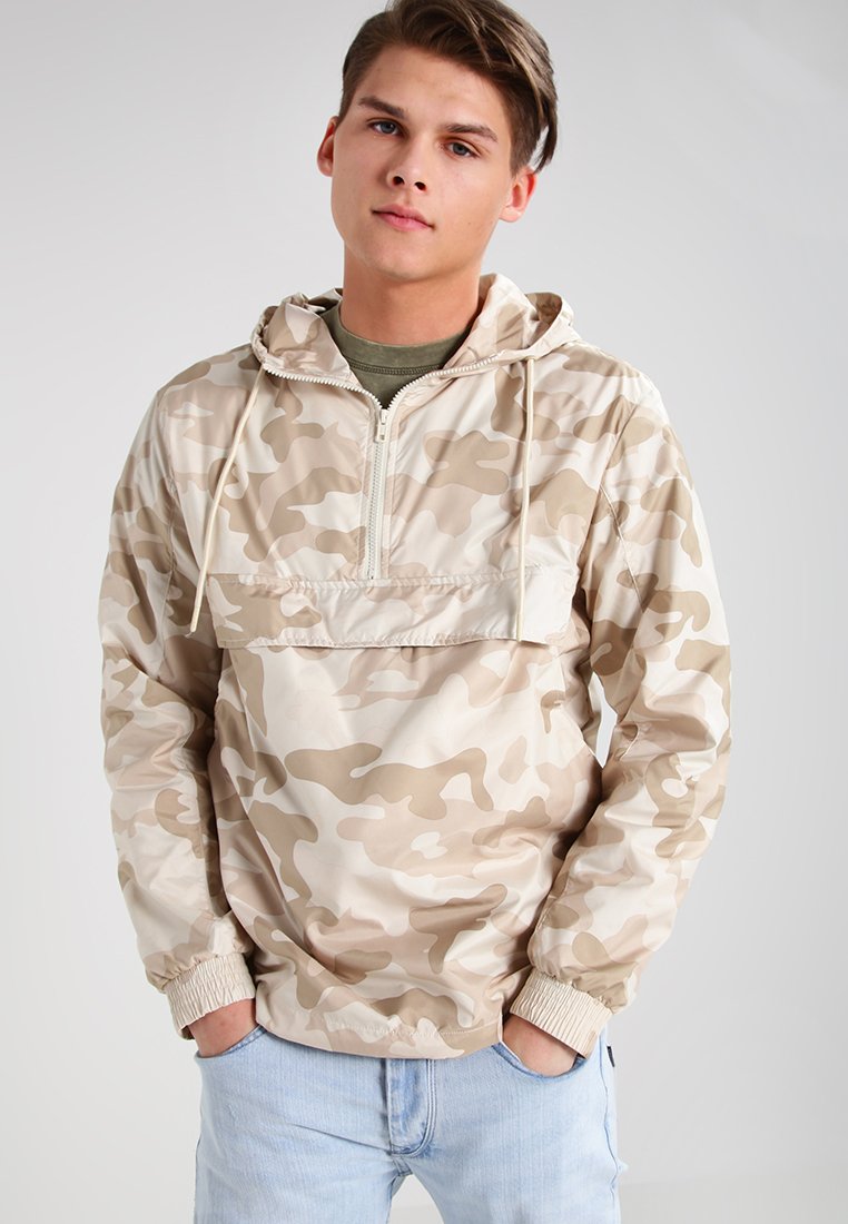 Chaqueta de media cremallera de camuflaje beige con capucha, puños elásticos y un bolsillo frontal; textura suave y material ligero.