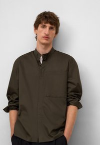 Donkergroene shirt met een hoge kraag, knoopsluiting aan de voorkant en een enige borstzak. Lange mouwen zijn opgerold bij de polsen. Glad stoftextuur.