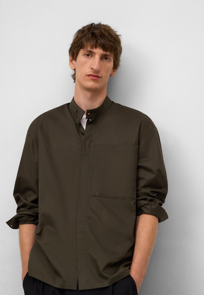 Donkergroene shirt met een hoge kraag, knoopsluiting aan de voorkant en een enige borstzak. Lange mouwen zijn opgerold bij de polsen. Glad stoftextuur.