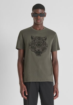Camiseta de algodón verde oliva con diseño de cabeza de tigre bordado en negro en la parte frontal, cuello redondo y mangas cortas.