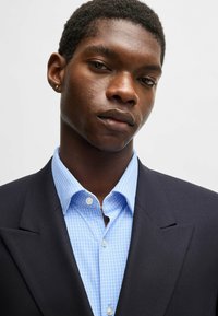 Blazer navy con rever classico sopra una camicia azzurro chiaro con un motivo geometrico e bottoni bianchi, che mostra una vestibilità strutturata.