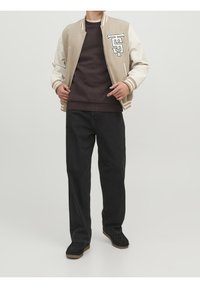 Giacca varsity beige con maniche crema, lettering ricamato sul petto, indossata sopra un maglione scuro e pantaloni neri, abbinata a scarpe scure.