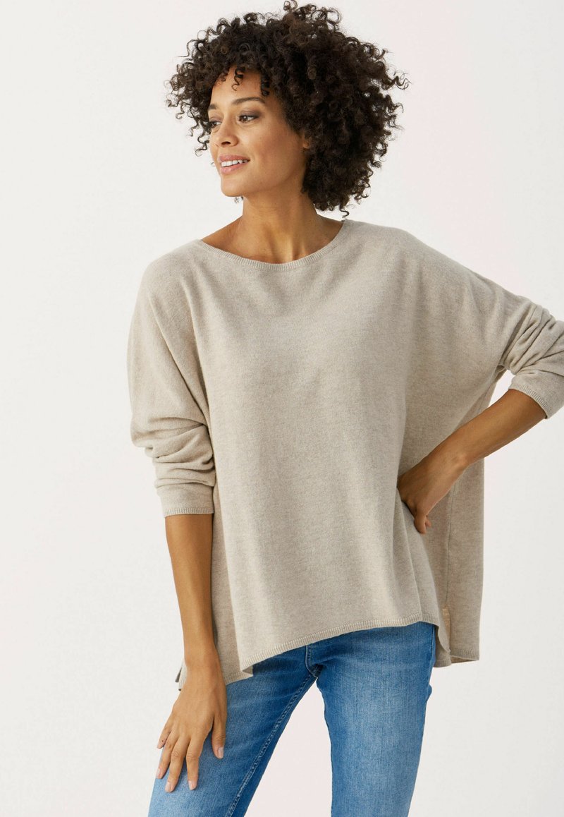 Hellbeige Oversize-Pullover mit weitem rundem Ausschnitt, langen Ärmeln und seitlichen Schlitzen, kombiniert mit blauen Jeans. Weiche Stricktextur.