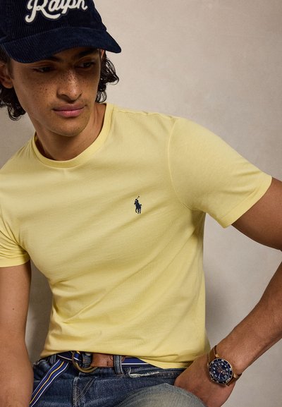 Mlad moški z pegami, ki nosi rumeno polo majico, mornarsko kapa s napisom "Ralph", modre kavbojke, črtast pas in uro, gleda navzdol.