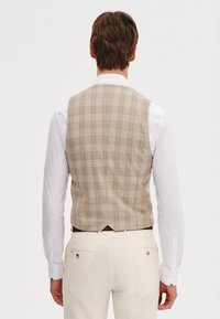 Beżowa kamizelka w kratę o dopasowanym kroju, wykonana z teksturowanej tkaniny; zestawiona z białą koszulą z długim rękawem i jasnymi spodniami.