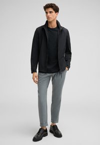 Schwarze Zip-Jacke mit hohem Kragen, kombiniert mit einem dunklen T-Shirt, hellgrauen schmal geschnittenen Hosen und schwarzen Schnürschuhen.