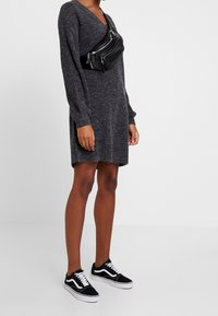 Grå stickad klänning med långa ärmar och V-ringning, tillsammans med en svart axelremsväska med flera fickor med dragkedja och svarta sneakers.