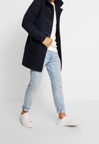 Granatowy płaszcz wierzchni z zapięciami na pętelki, boczne kieszenie, w połączeniu z jasnoniebieskimi podwiniętymi dżinsami i białymi trampkami, prezentujący casualowy styl.