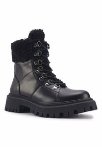 Botas de cuero negras hasta el tobillo con una suela gruesa y acanalada, cuello forrado de piel y ojetes metálicos para los cordones. Presentan una textura lisa y un diseño robusto.