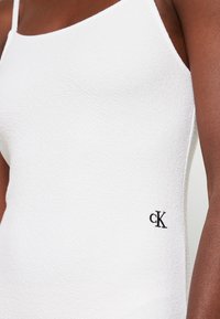 Biała bezrękawnikowa odzież z teksturowanej tkaniny, z czarnym logo "ck" w dolnym rogu. Cienkie ramiączka i dopasowany fason.