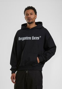 Sweatshirt à capuche noire avec un graphique blanc "visages oubliés", poche kangourou et poignets côtelés. Texture douce et coupe décontractée.
