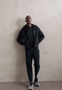 Sudadera con cremallera negra con un patrón, encima de una camiseta negra, combinada con pantalones negros que tienen una franja blanca a los lados y zapatillas gris. Textura suave.