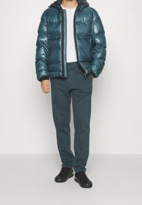 Teal pufferjacka med glänsande yta, zip-stängning och huva, kombinerad med mörkgröna byxor och svarta sneakers. En enkel vit T-shirt under.