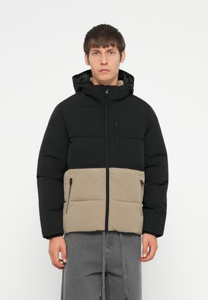 Chaqueta tipo puffer con la parte superior negra y la inferior beige, que cuenta con un cierre frontal, bolsillos laterales y una capucha. Textura suave.