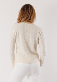 Donna con i capelli lunghi che indossa un maglione cream a trecce e pantaloni bianchi, voltata verso l'indietro su uno sfondo chiaro e semplice.