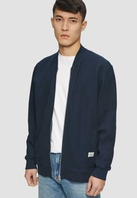 Veste bomber zippée bleu marine avec des poignets côtelés, texture lisse, design non structuré, portée avec un t-shirt blanc et un jean bleu.