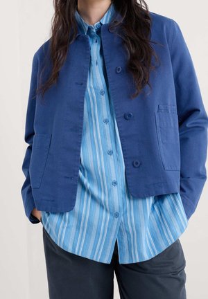 Femme portant une veste bleu foncé par-dessus une chemise bleu clair à rayures verticales, les mains dans les poches, pantalon foncé visible.